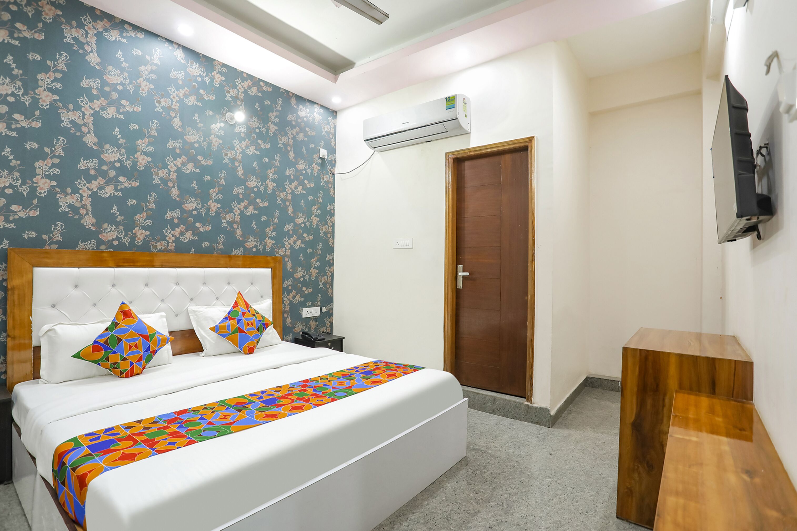 A bedroom with a bed, a wooden headboard, a floral-patterned wall, an air conditioning unit, a wooden door, and a small wooden table.