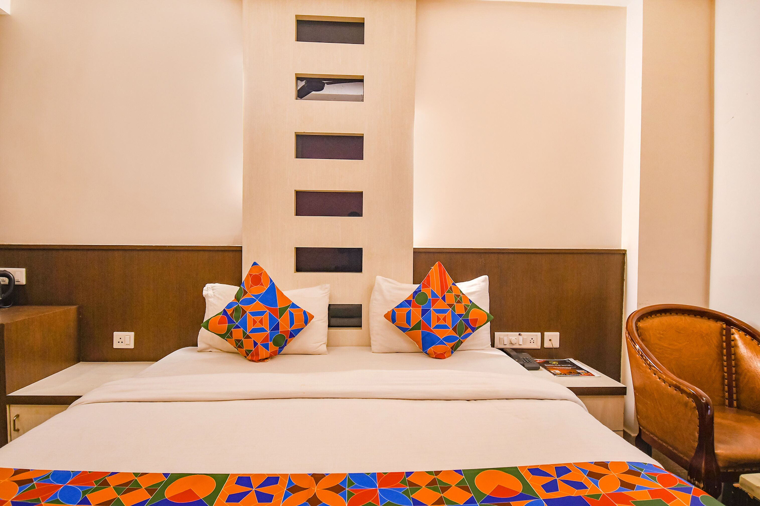 Une chambre d’hôtel comprenant un lit, deux oreillers à motifs géométriques, une tête de lit en bois, une table de chevet, une chaise et un mur orné d’un panneau vertical.