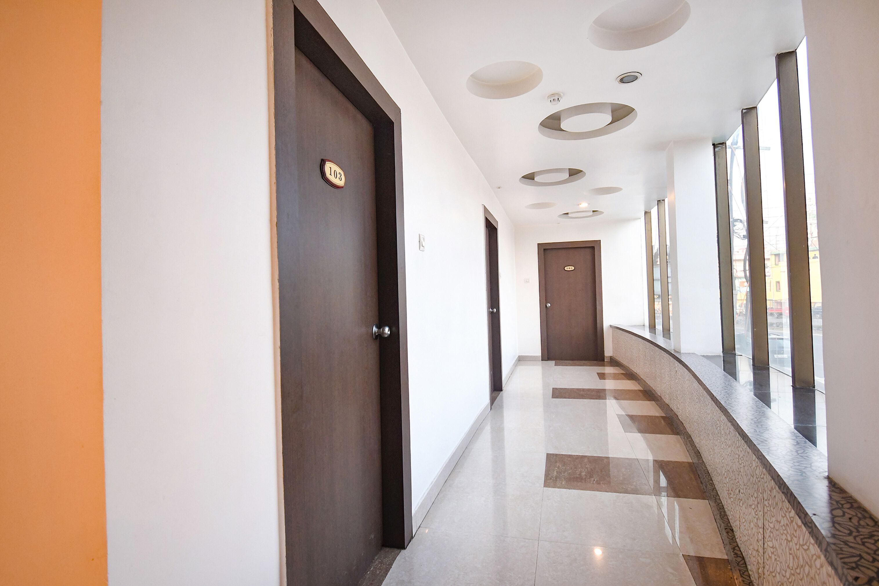 Un pasillo moderno de hotel con piso a cuadros, una puerta de madera con placa de número y un muro de vidrio con ventanas.