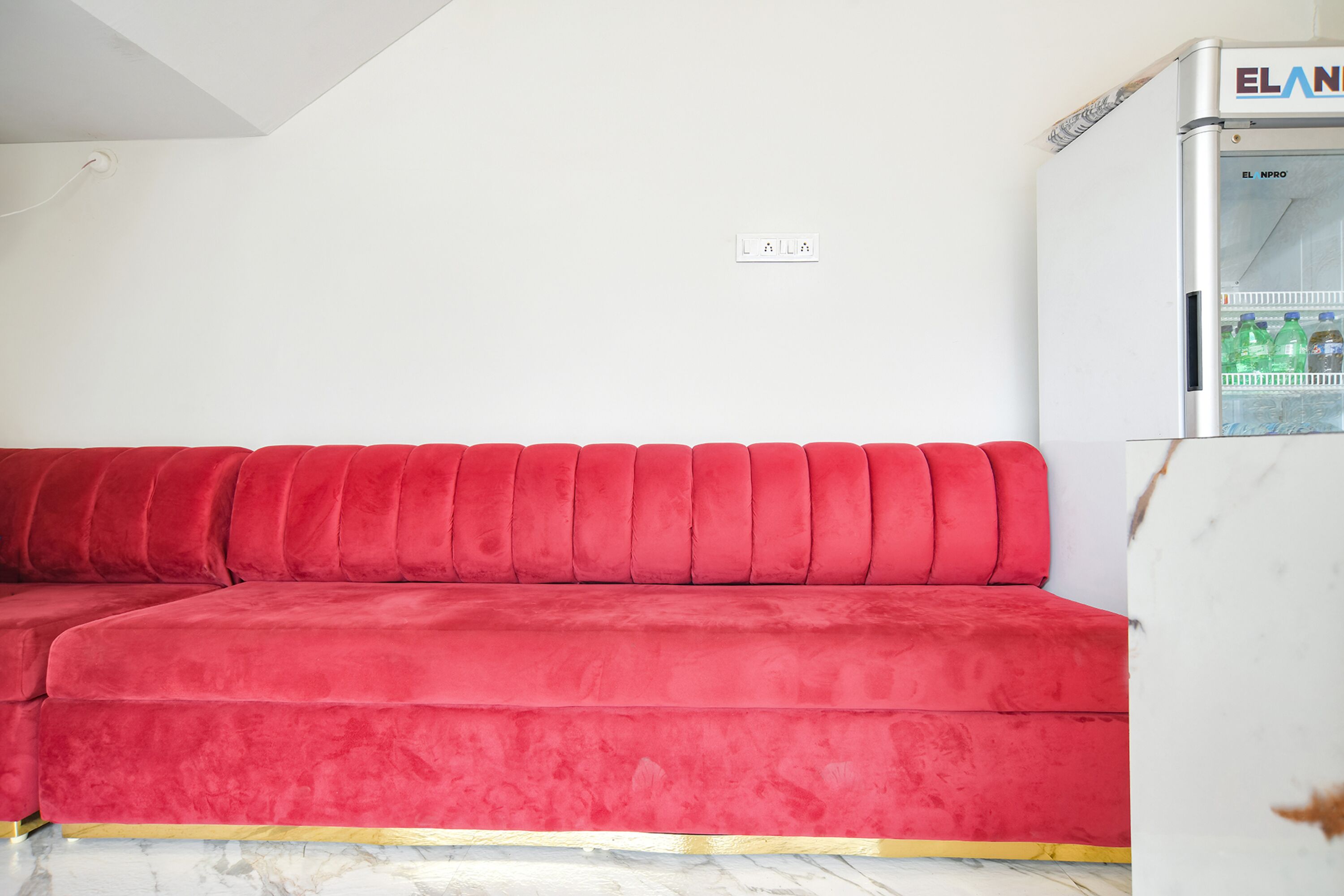 A red sofa in a room with a white wall and a white refrigerator with a water dispenser.
