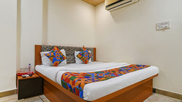 A neatly made bed with a colorful geometric-patterned blanket, a wooden headboard, and a bedside table with a remote control and a water bottle. - Fabexpress Brahmasmi (Ujjain)