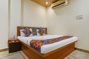 A neatly made bed with a colorful geometric-patterned blanket, a wooden headboard, and a bedside table with a remote control and a water bottle. - Fabexpress Brahmasmi (Ujjain)