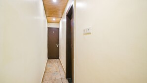 A narrow hallway with a wooden ceiling, a brown door at the end, and a light switch on the wall. - Fabexpress Brahmasmi (Ujjain)