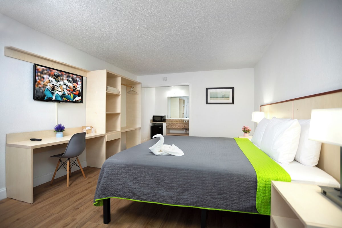 A hotel room with a bed, a desk, a chair, a television, a mirror, and a nightstand.