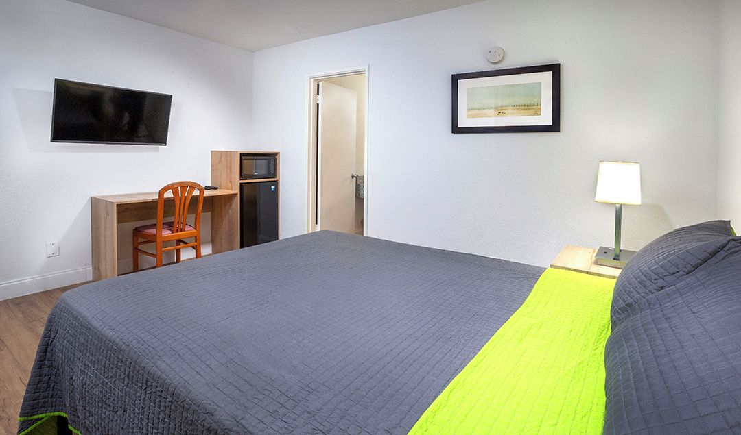 A hotel room with a bed, a desk with a chair, a television, a door, and a framed picture on the wall.