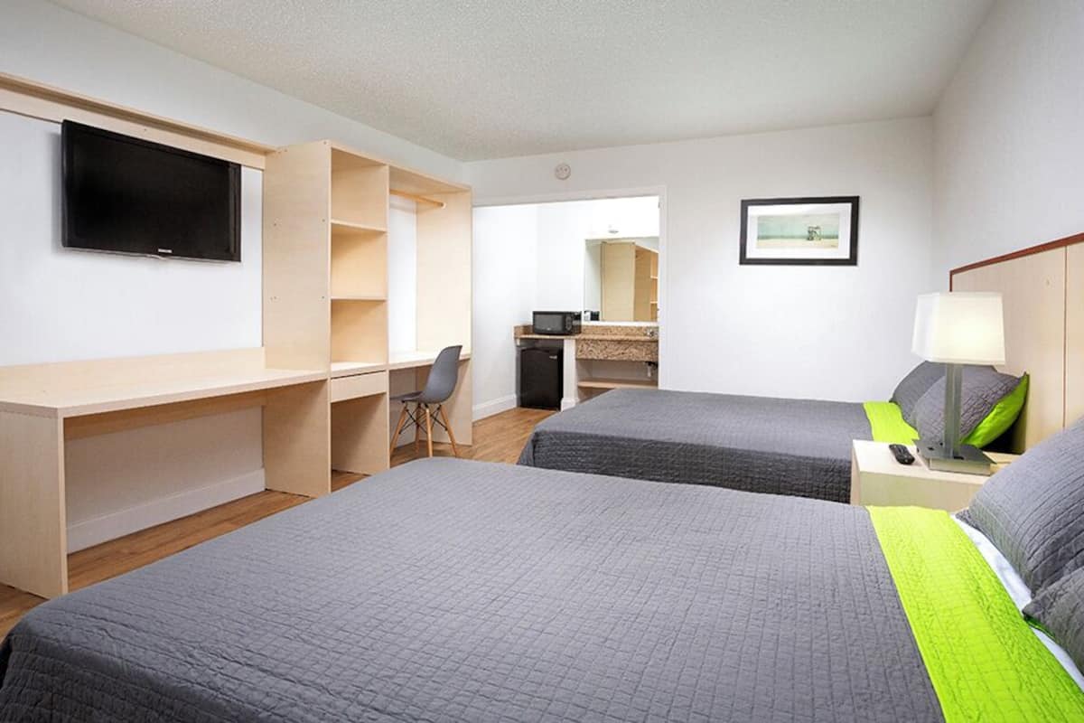 A hotel room with two beds, a desk, a TV, a mirror, and a small kitchenette.