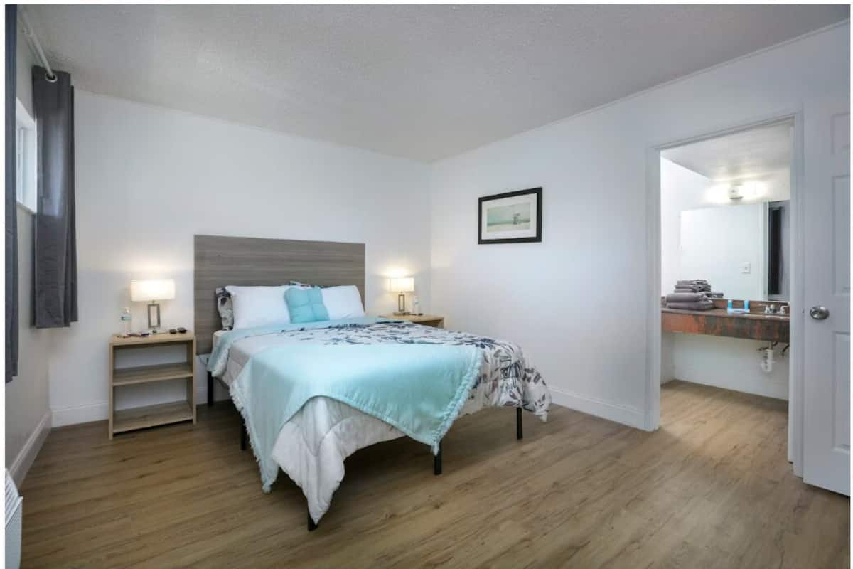 A bedroom with a bed, bedside tables, a nightstand, a framed picture, and a bathroom visible through an open door.