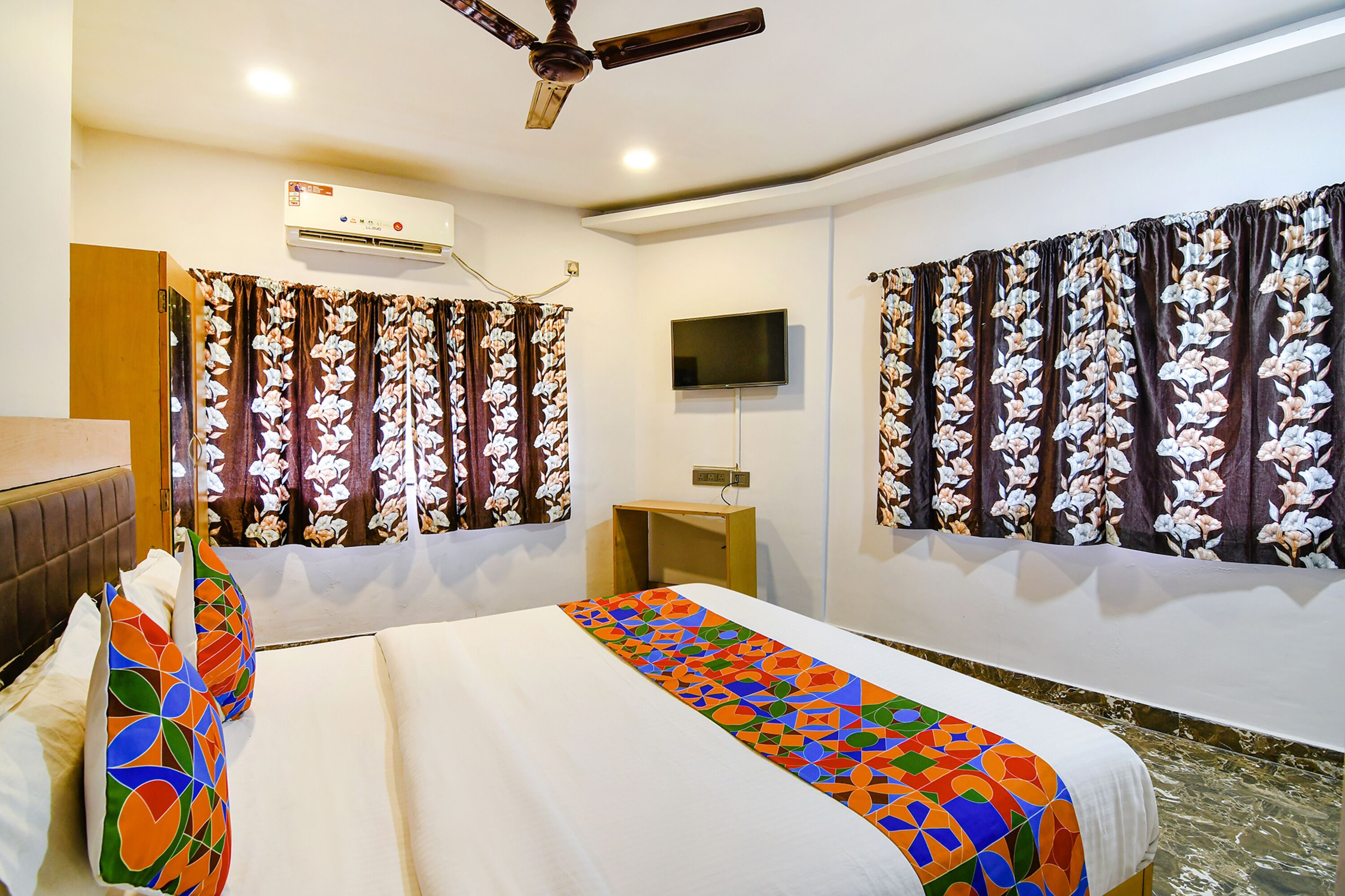 Une chambre d’hôtel équipée d’un lit, d’une télévision, d’un climatiseur et de rideaux à motifs floraux.