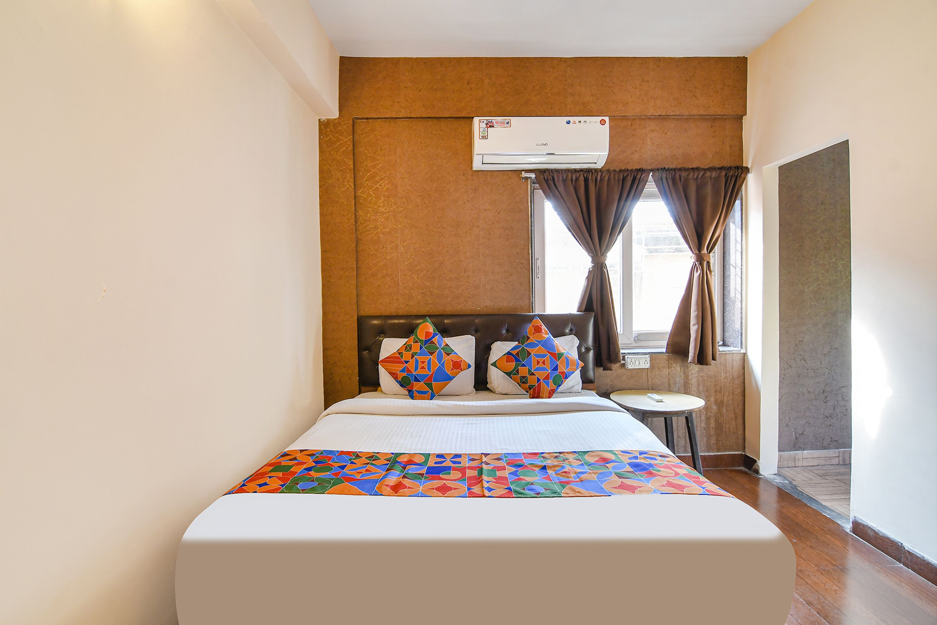 A hotel room with a bed, a small table, an air conditioning unit, and a window with curtains.