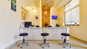 A reception area with a wooden panel wall, a reception desk, three black office chairs, a television on the wall, and a window with blinds. - Fabhotel Aayojan (Jaipur)