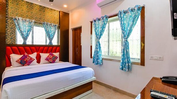 A hotel room with a red upholstered headboard, a bed with white bedding and a blue accent, two windows with blue curtains, and a TV on the wall. - Fabexpress Golden Palace (Kolkata)