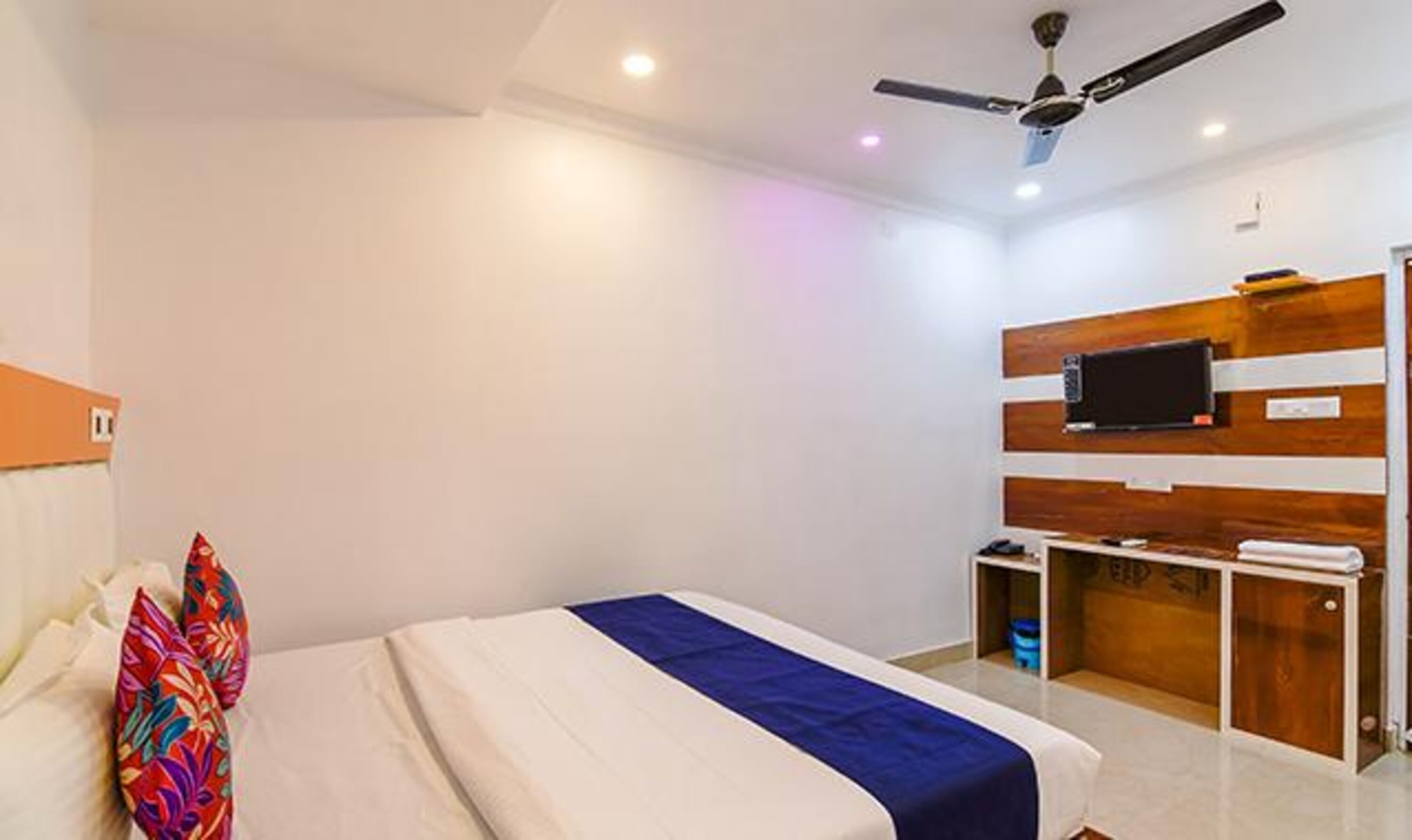 A bedroom with a bed, a ceiling fan, a TV mounted on the wall, and a small desk with a chair.