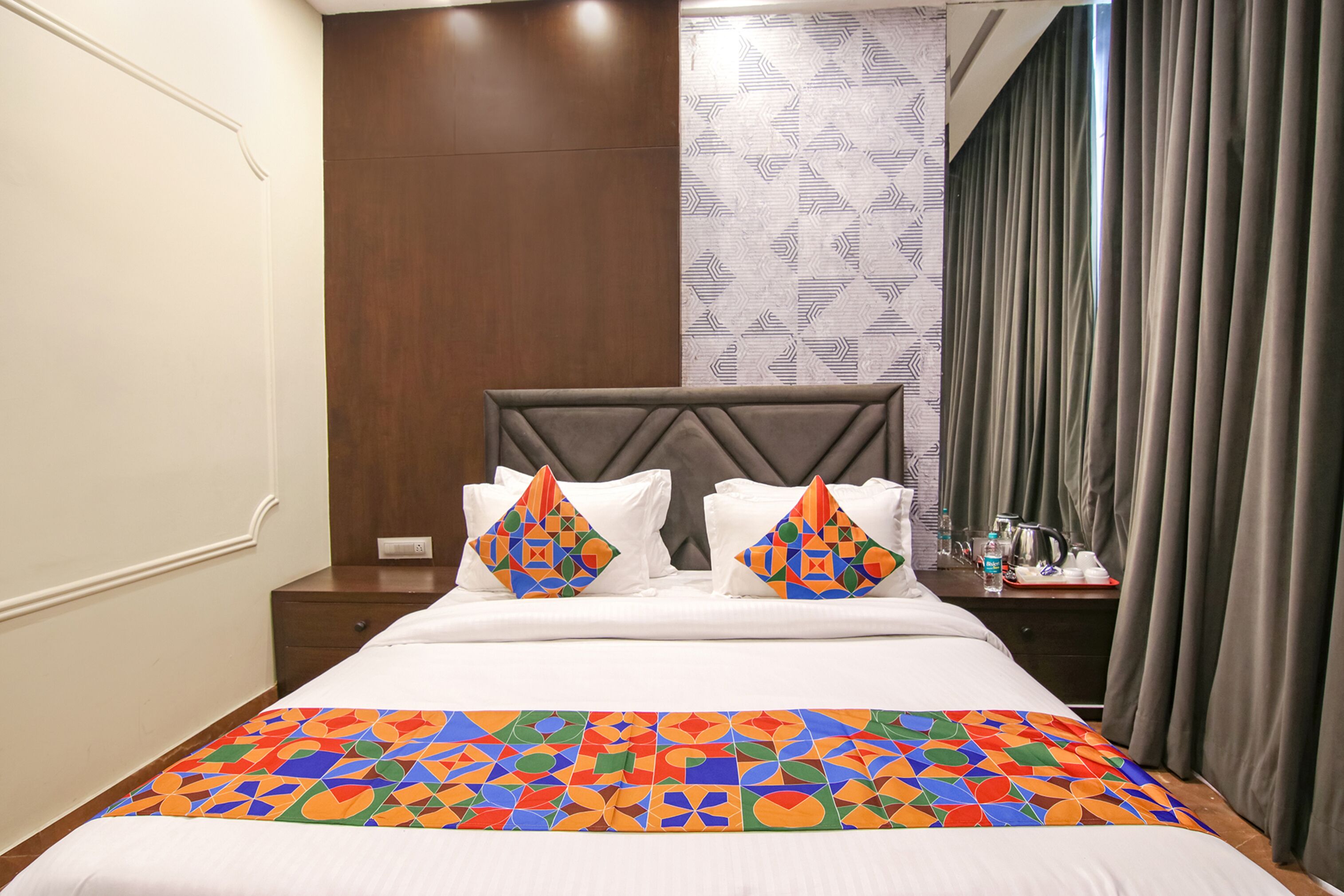 A hotel room with a large bed, two pillows, a bedside table, a nightstand, a window with curtains, and a patterned wall.