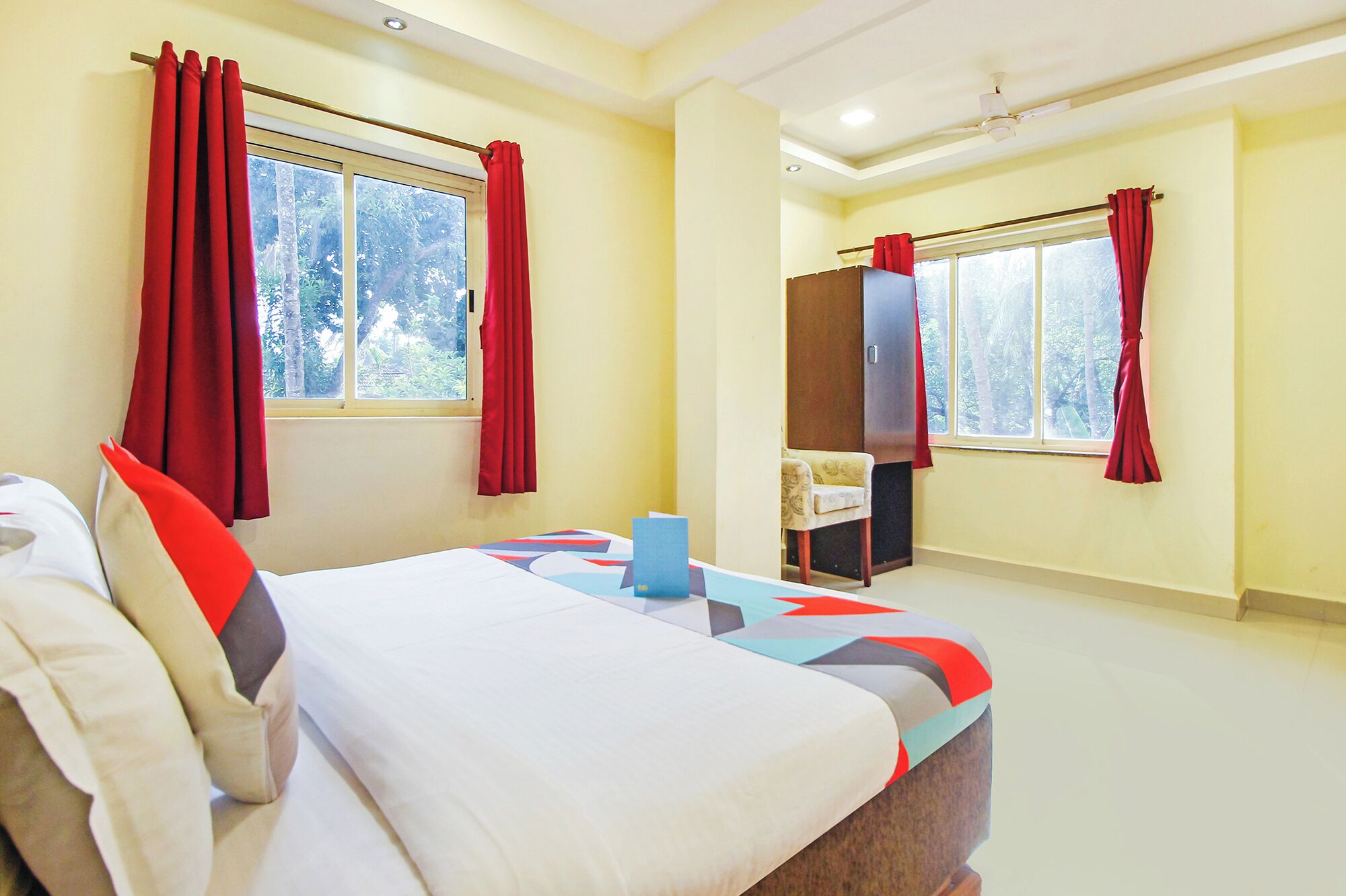 A hotel room with a bed, two windows with red curtains, a chair, and a desk.