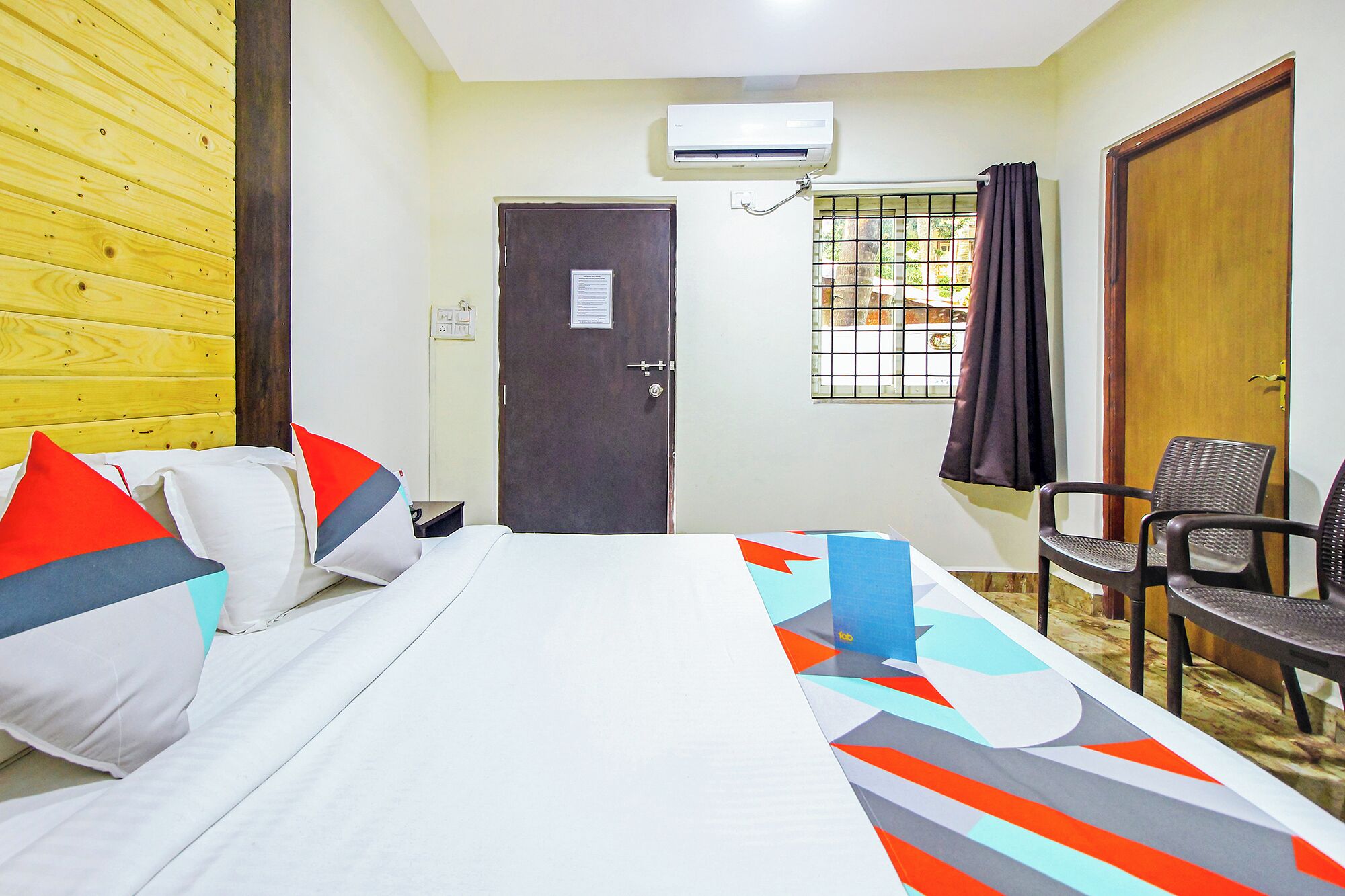 A hotel room with a bed, a chair, an air conditioning unit, a door, and a window with a curtain.