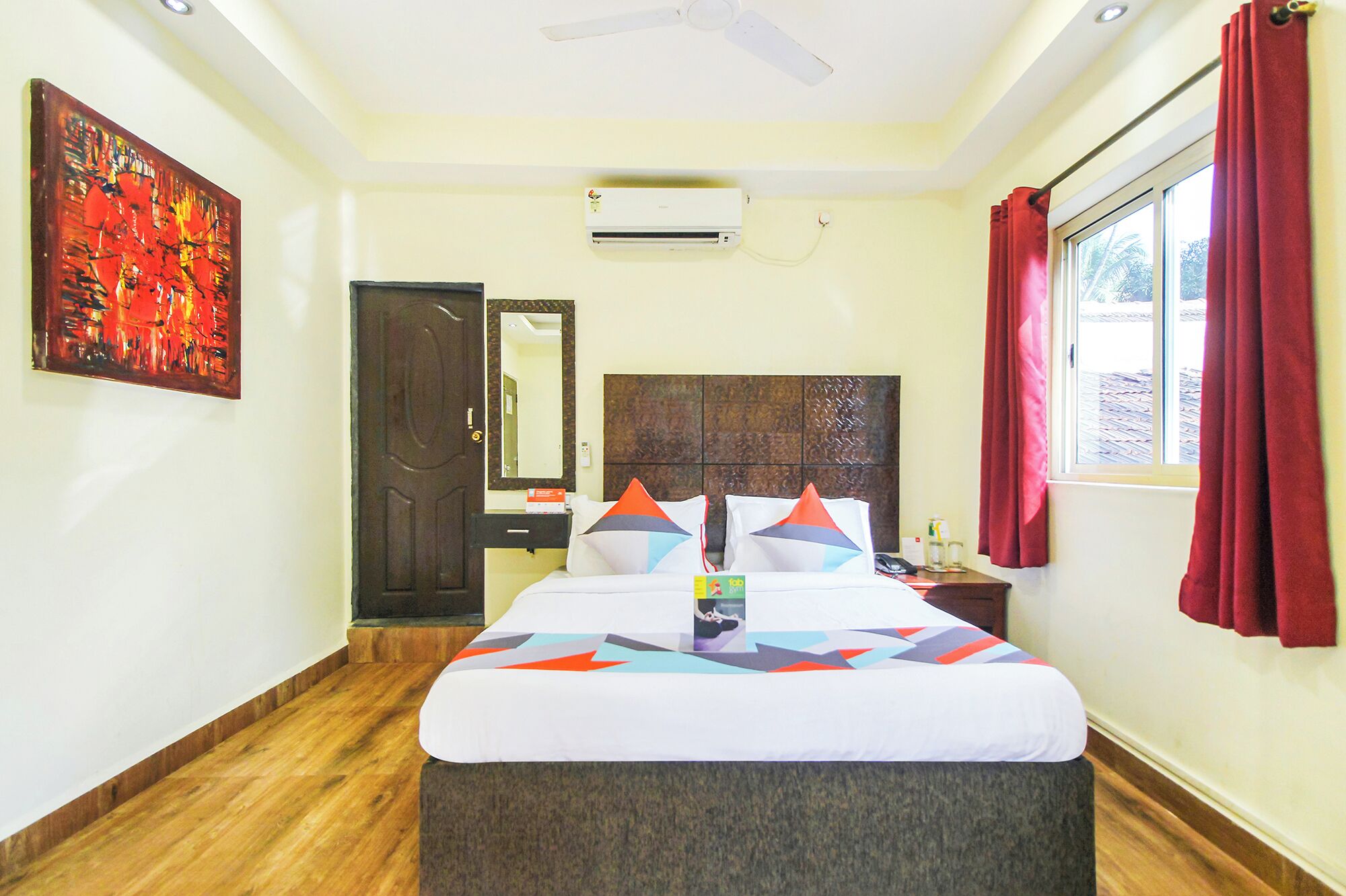 A hotel room with a bed, a nightstand, a mirror, a window with red curtains, and an air conditioning unit.