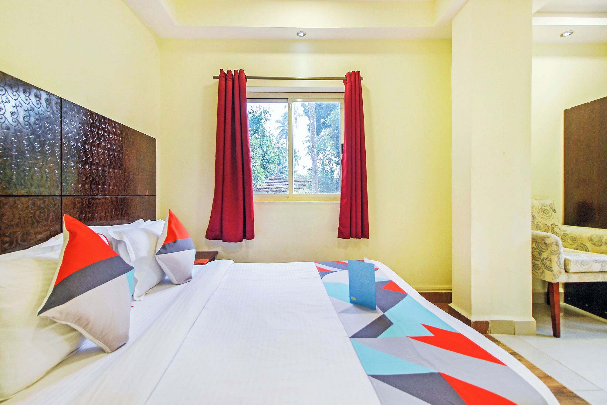 A hotel room with a bed, a window with red curtains, and a chair in the corner.