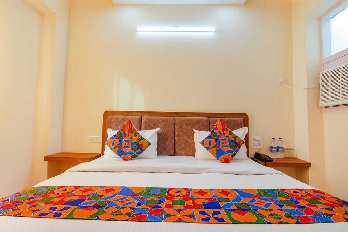 A hotel room with a bed, two pillows, a bedside table with a phone, and a window with blinds.