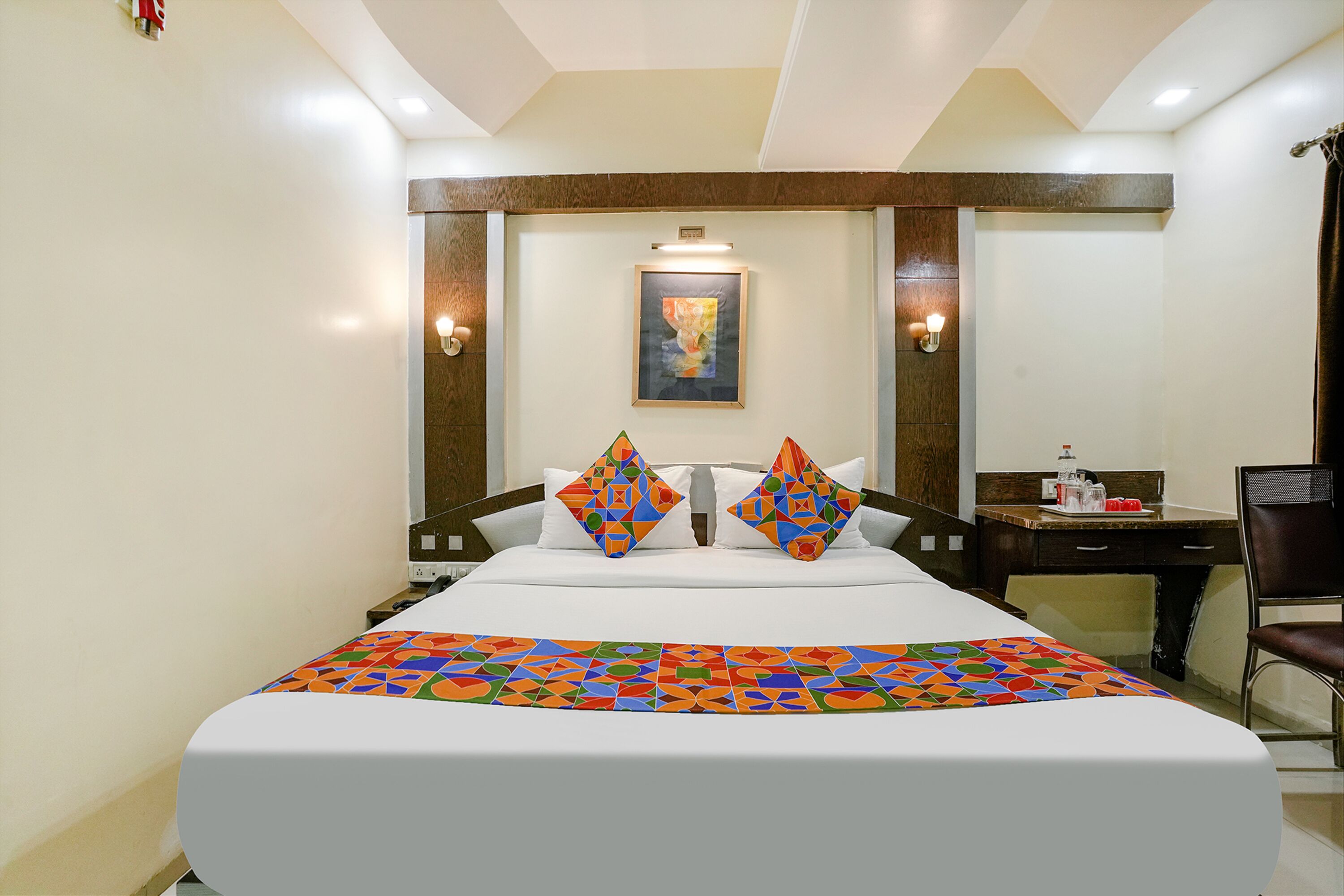 A hotel room with a large bed, two decorative pillows, a painting on the wall, and a desk with a chair.