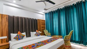 A hotel room with a bed, a ceiling fan, teal curtains, and a woven chair. - Fabhotel Ayana Inn (Dharamshala)