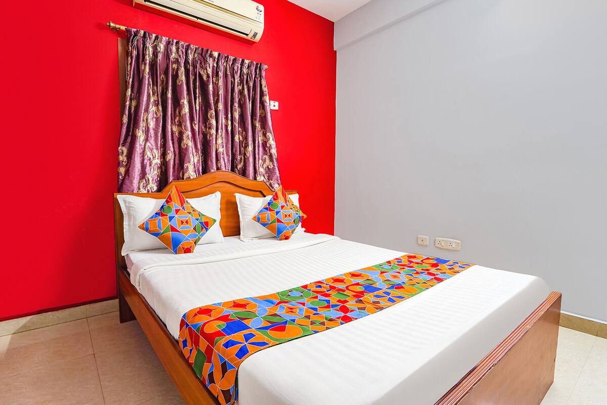 A hotel room with a bed, a red wall, a window with curtains, and an air conditioning unit.