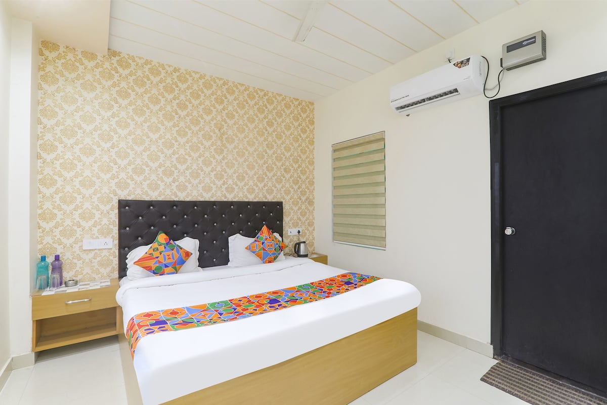 A hotel room with a bed, a nightstand, a window with blinds, an air conditioning unit, and a door.
