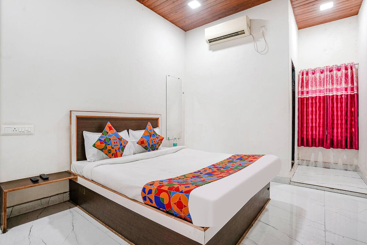 A hotel room with a bed, a nightstand, an air conditioning unit, and a window with curtains.