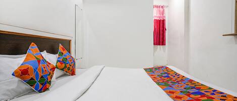 A hotel room with a bed, colorful pillows, a patterned bed runner, a window with curtains, and an air conditioning unit.