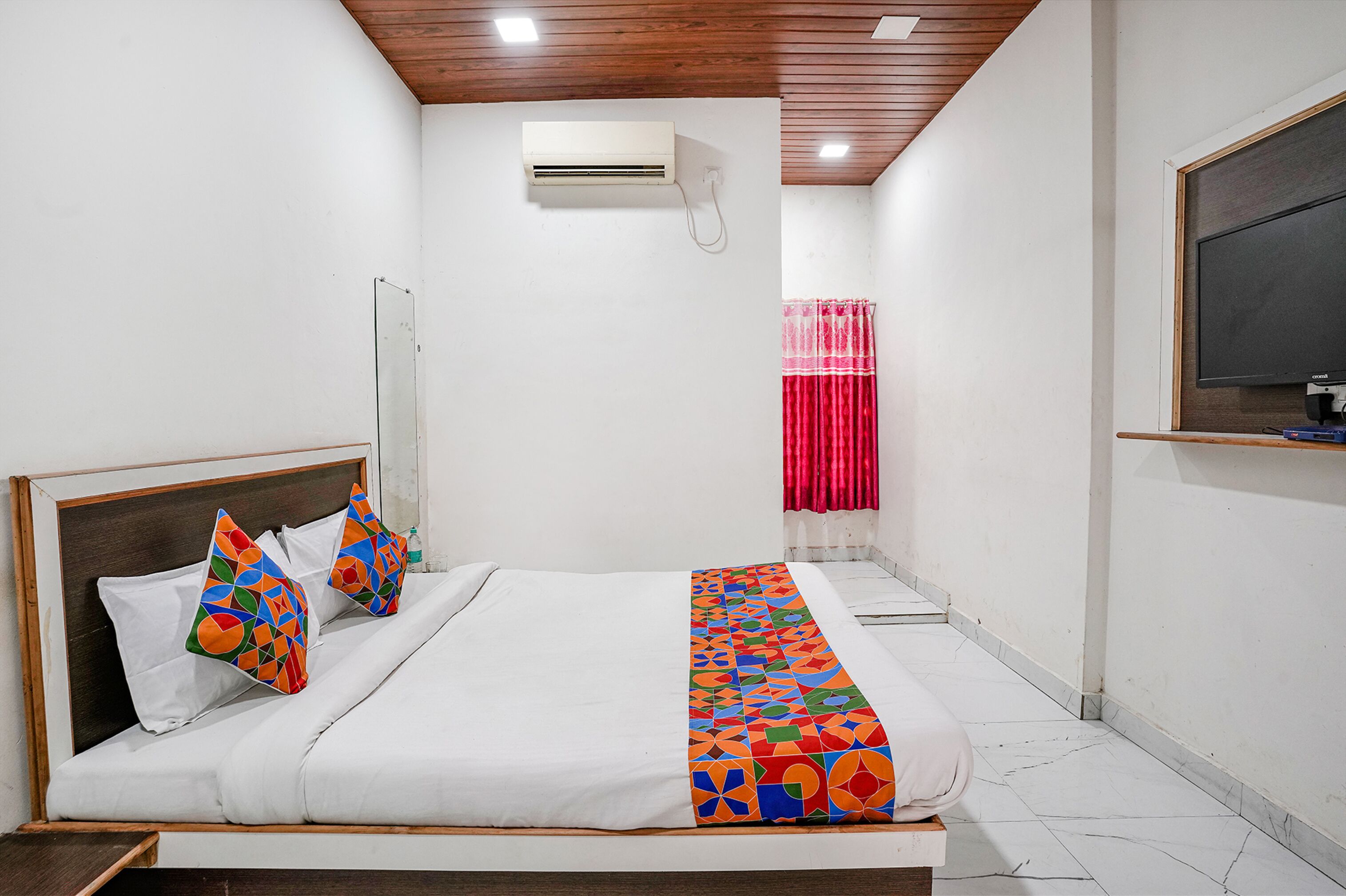 Habitación de hotel con cama, techo de madera, unidad de aire acondicionado, cortina roja y televisión montada.