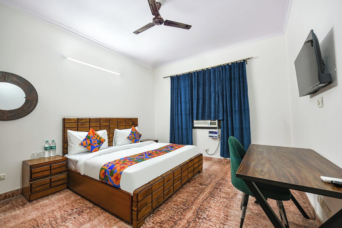 Une chambre d’hôtel avec un lit en bois, un bureau, une chaise, une télévision, un ventilateur de plafond et une fenêtre avec des rideaux bleus.