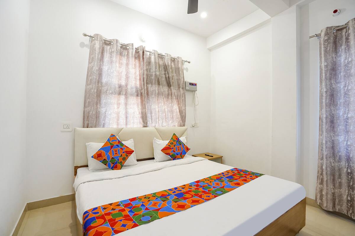 A hotel room with a bed, white curtains, a heater, and a power outlet.