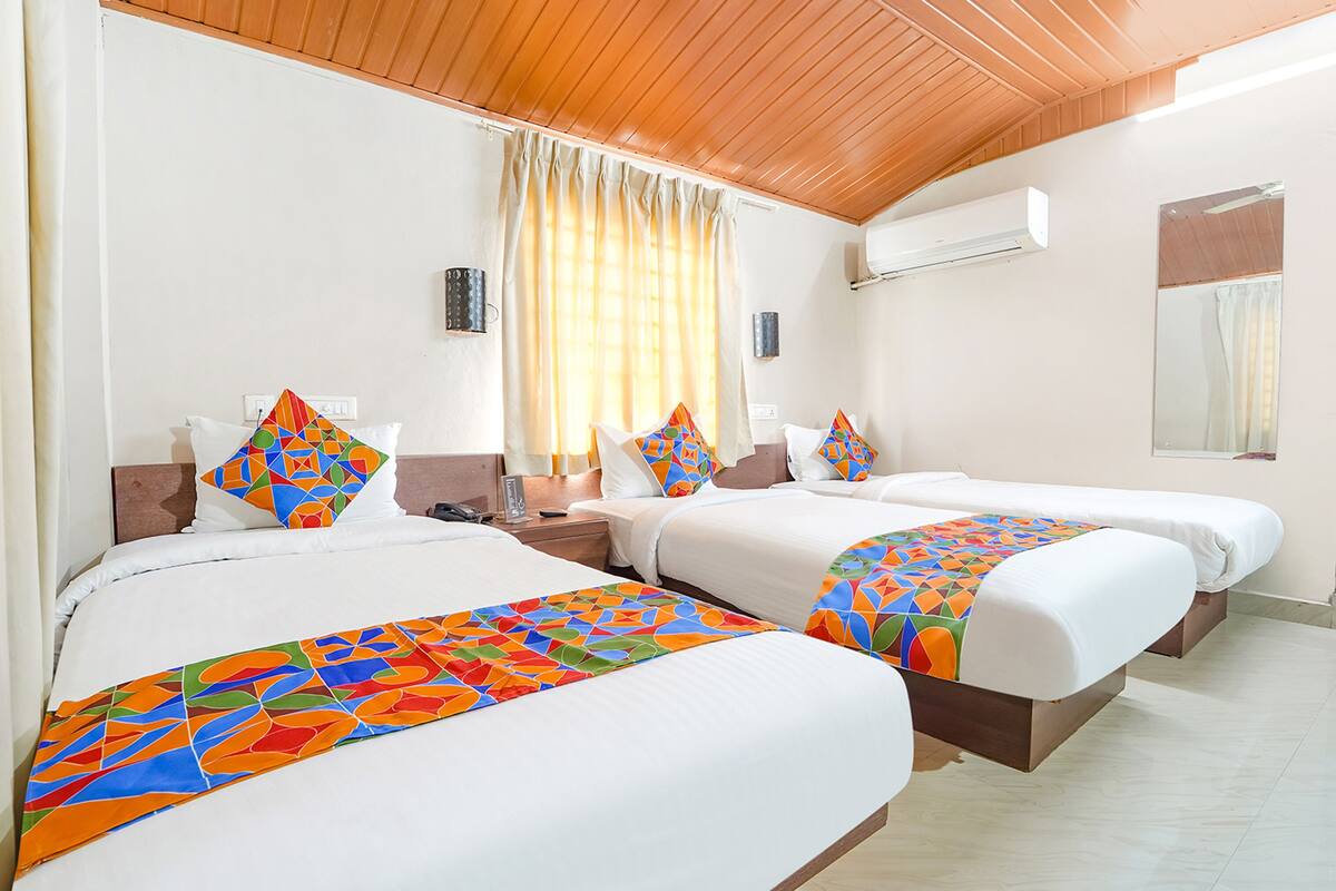 Ein Hotelzimmer mit zwei Betten, jeweils mit farbenfrohen, geometrischen Tagesdecken und passenden Kissen. Es gibt eine Holzbalkendecke, eine Klimaanlage und ein Fenster mit Vorhängen.