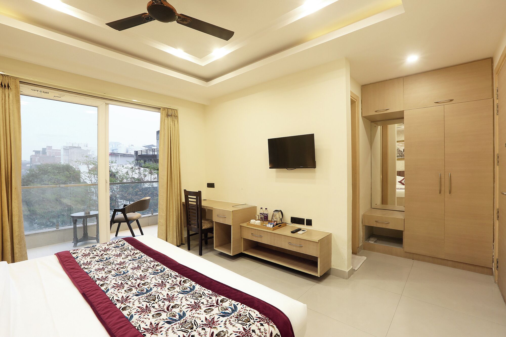 A hotel room with a bed, a desk, a chair, a TV, a wardrobe, and a balcony with a view of the city.