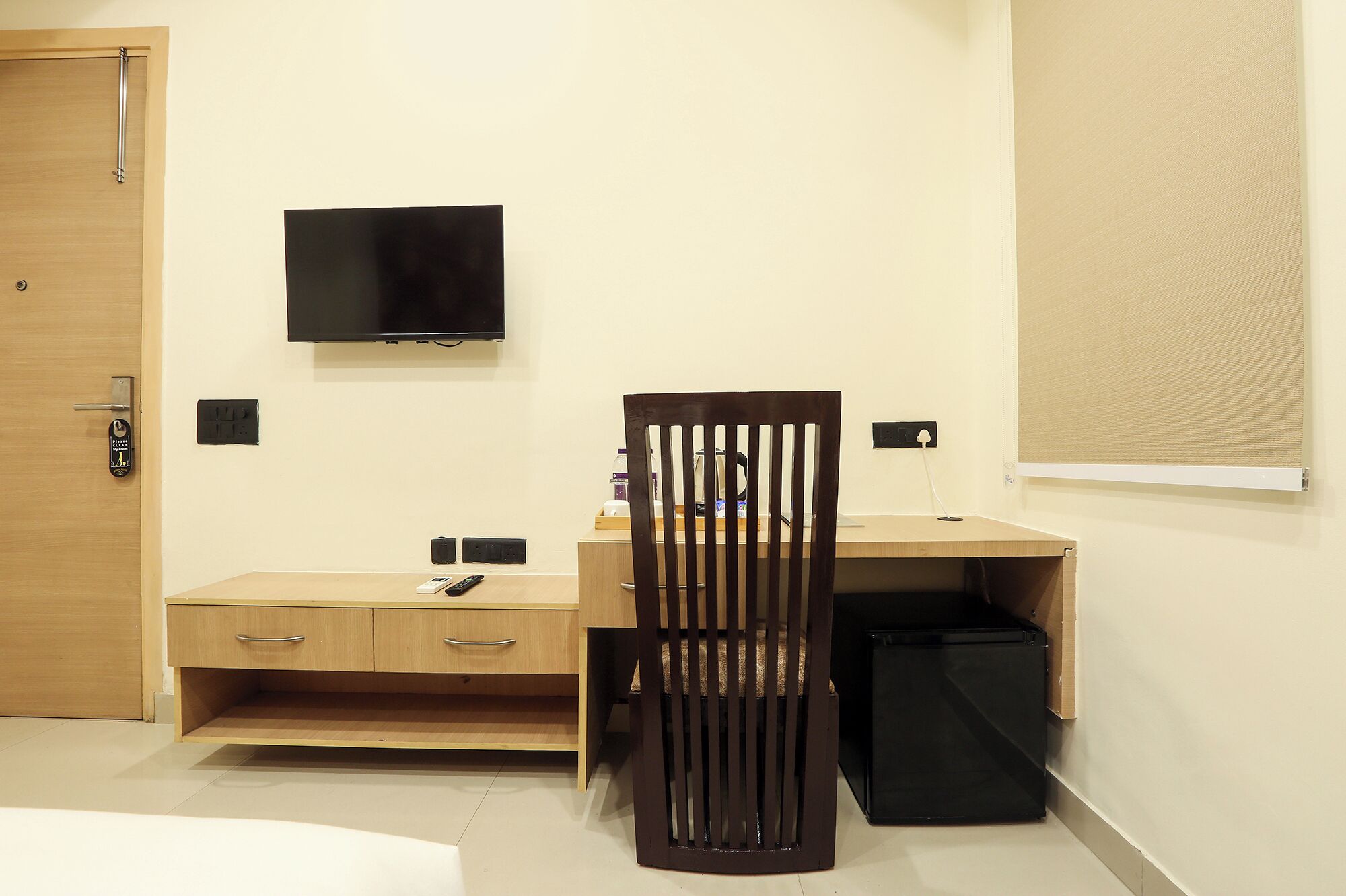 A hotel room with a wooden desk, a television mounted on the wall, a chair, a small black trash bin, and a window with a blind.