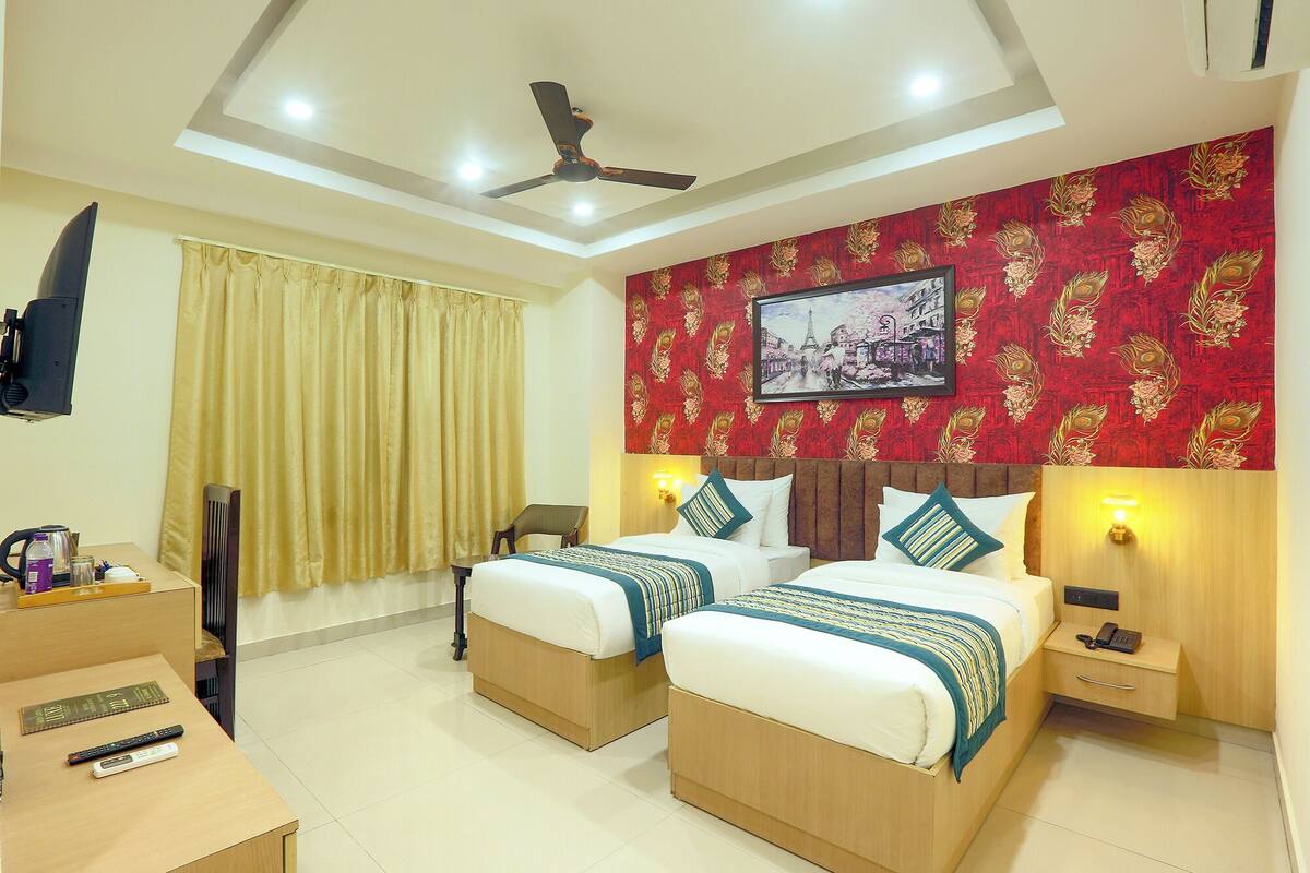 A hotel room with two beds, a ceiling fan, a TV, a desk with a kettle, and a telephone.