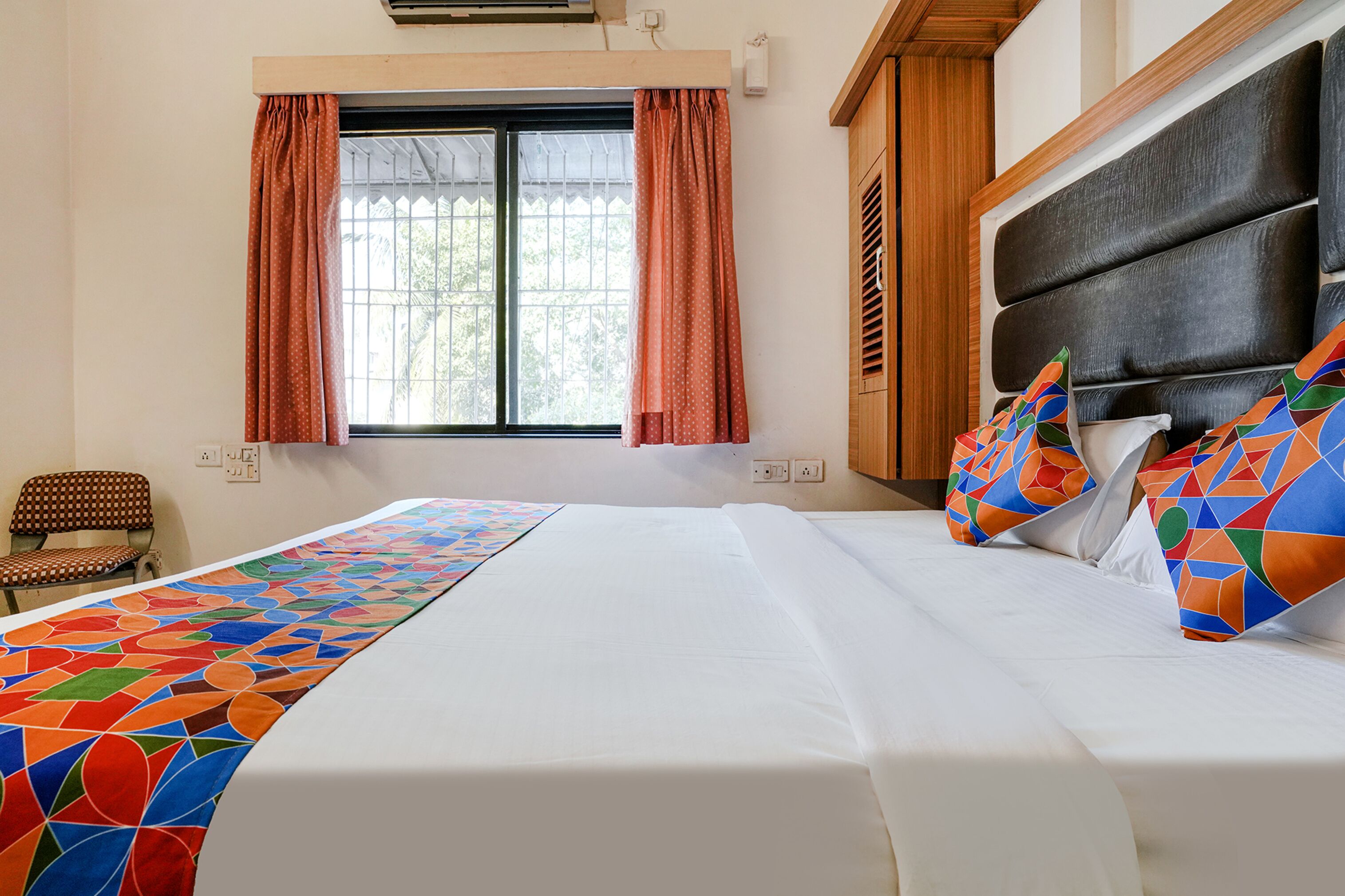 A hotel room with a bed, a window with curtains, a chair, and a headboard.