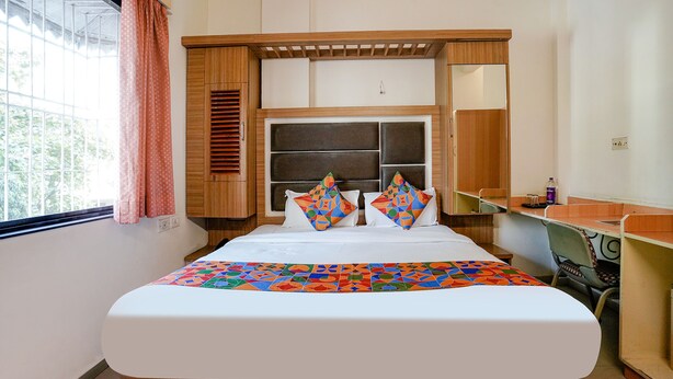 A hotel room with a bed, a wooden headboard, a desk, and a window with curtains. - Fabexpress Shanti Inn (Nashik)