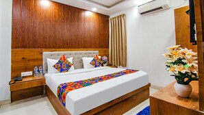 A hotel room with a wooden headboard, a bed with colorful bedding, a nightstand with a phone and water bottles, a vase with flowers, and an air conditioning unit on the wall. - Fabhotel The Glory I (New Delhi)