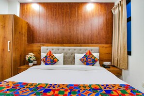 A bedroom with a tufted headboard, a bed with white bedding, two colorful geometric pillows, a small bedside table with a vase of flowers, and a wooden wardrobe. - Fabhotel The Glory I (New Delhi)
