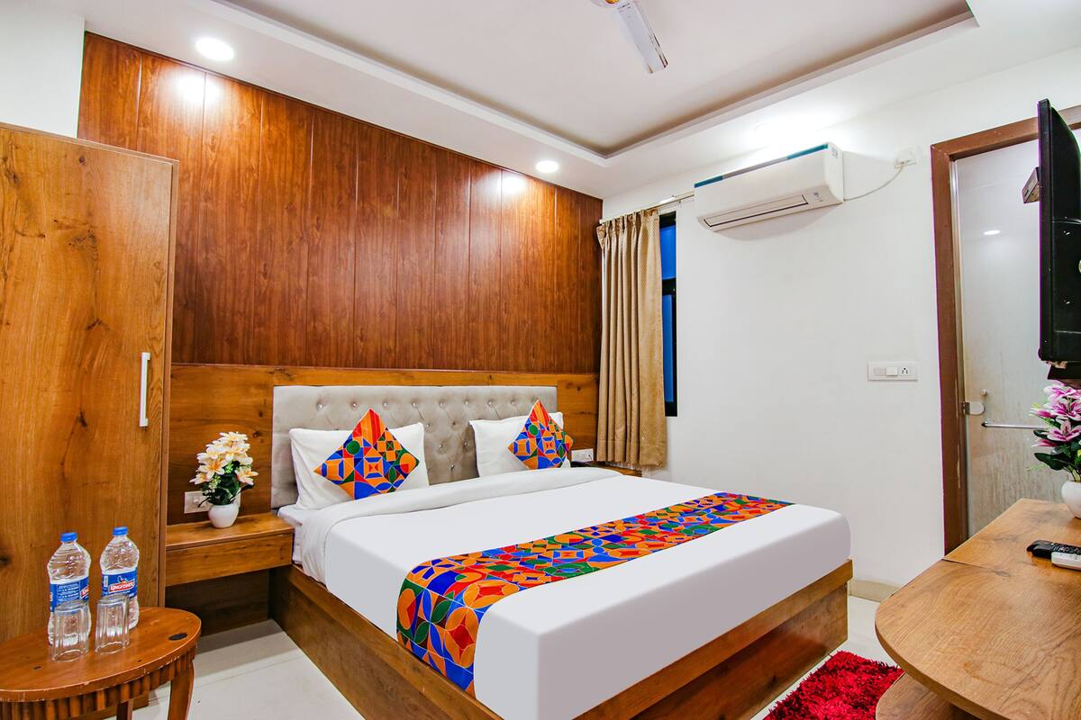 A hotel room with a wooden headboard, a bed with a colorful blanket, a nightstand with water bottles, a small table with a remote control, and an air conditioning unit on the wall.
