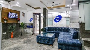 A reception area with a blue sofa, a glass coffee table, a TV mounted on a wooden panel, and a ceiling fan. - Fabhotel The Glory I (New Delhi)