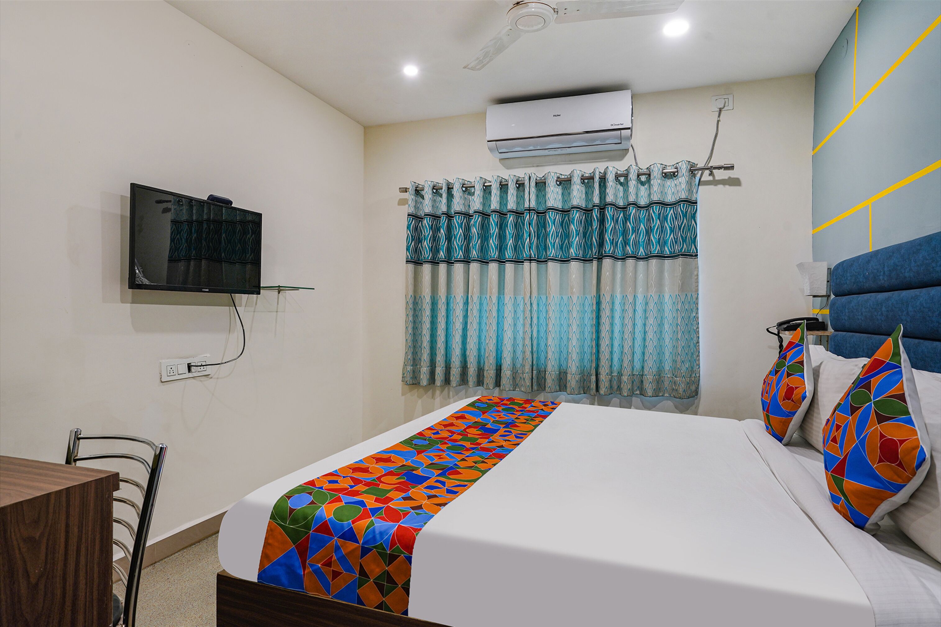 Quarto de hotel com cama, TV fixada na parede, ventilador de teto, aparelho de ar condicionado e janela com cortinas.