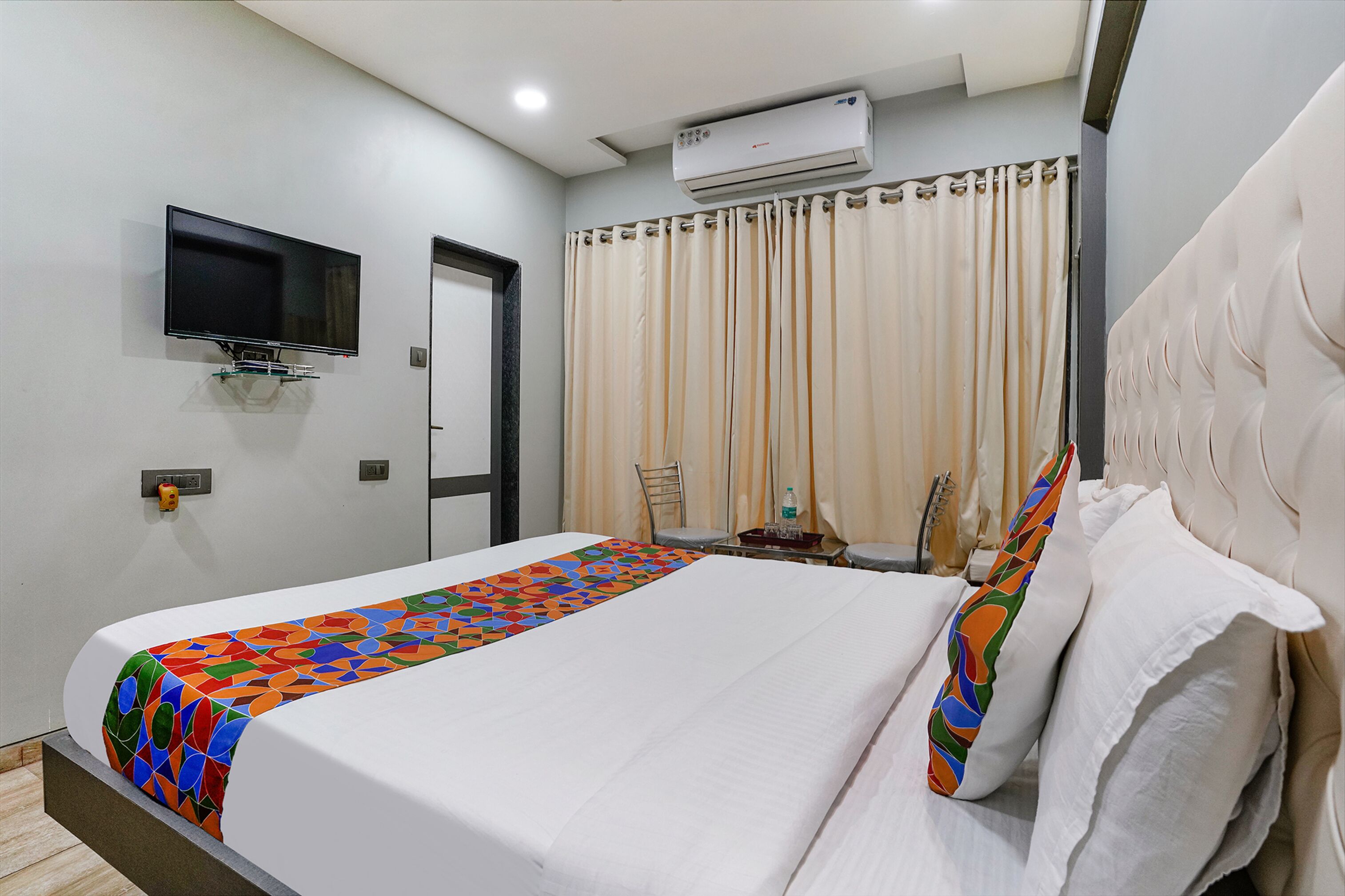 A hotel room with a bed, a TV mounted on the wall, an air conditioner, a curtain, and a small table with a bottle.
