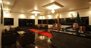 A modern living room with a dark marble floor, a central seating area with sofas and armchairs, a coffee table, and a red rug. There are large windows providing natural light and a view of the outside. - Fabhotel Prime Moon Hill Resorts (Devikolam)