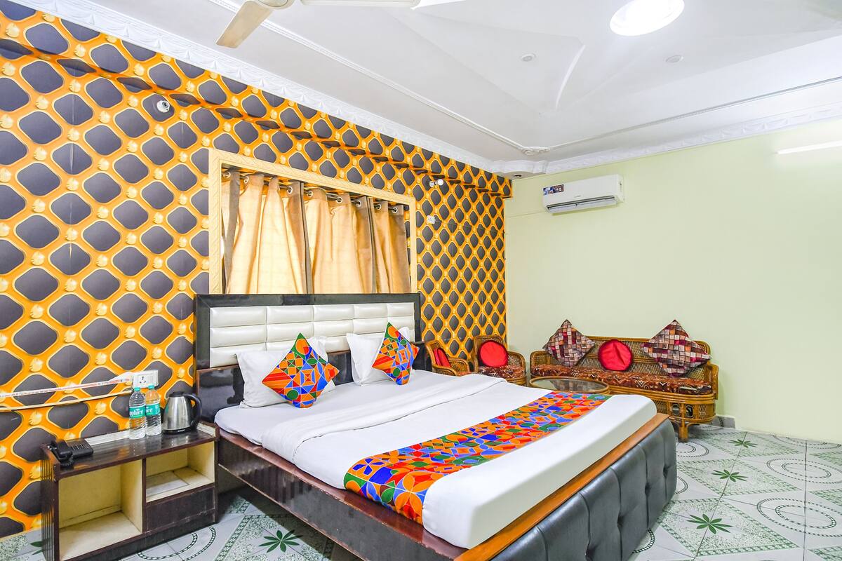 A hotel room with a patterned wall, a bed with a colorful bedspread, a bedside table with a phone and water bottles, and a wicker chair.