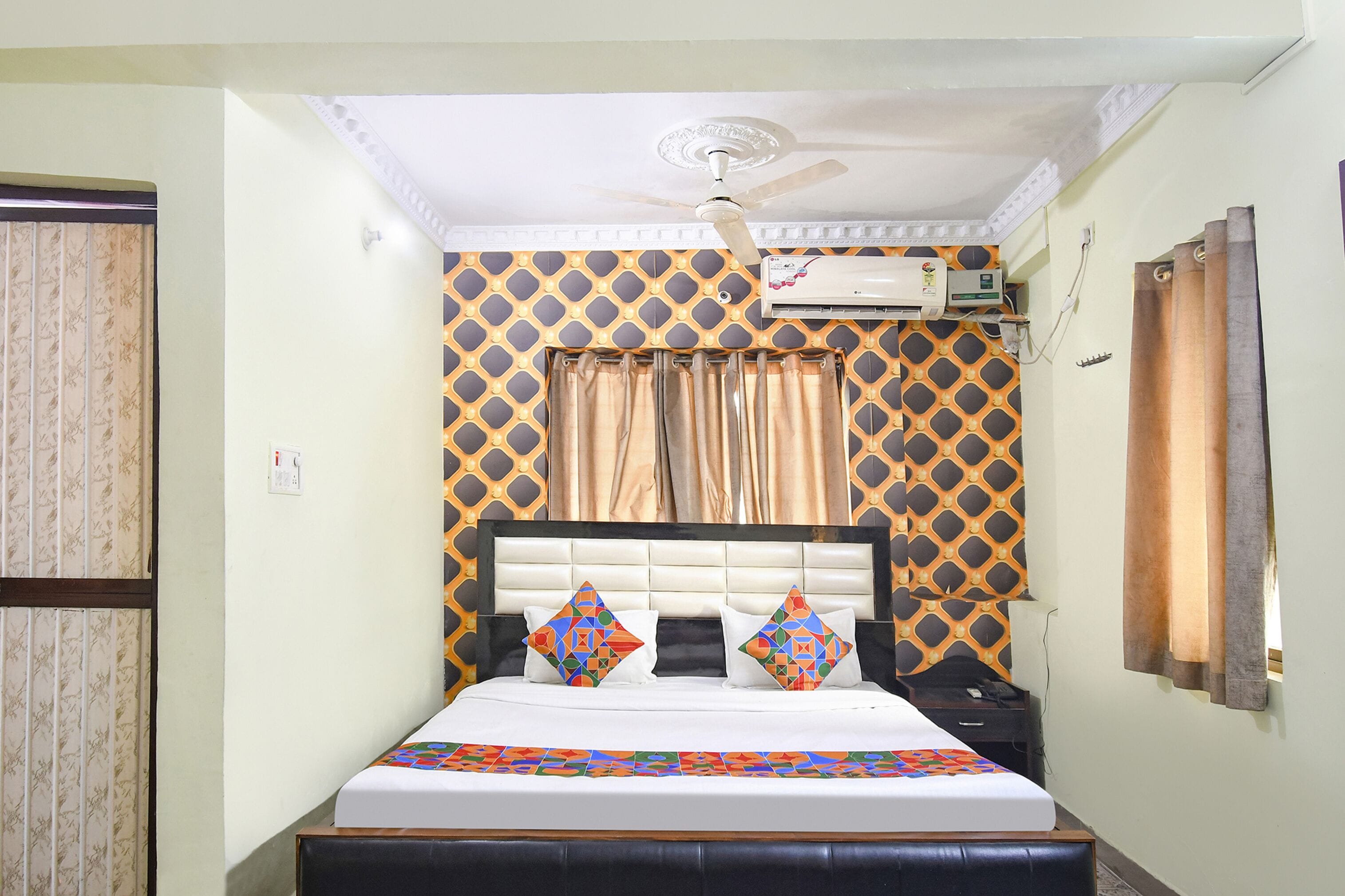 A hotel room with a bed, a ceiling fan, an air conditioning unit, and patterned curtains.