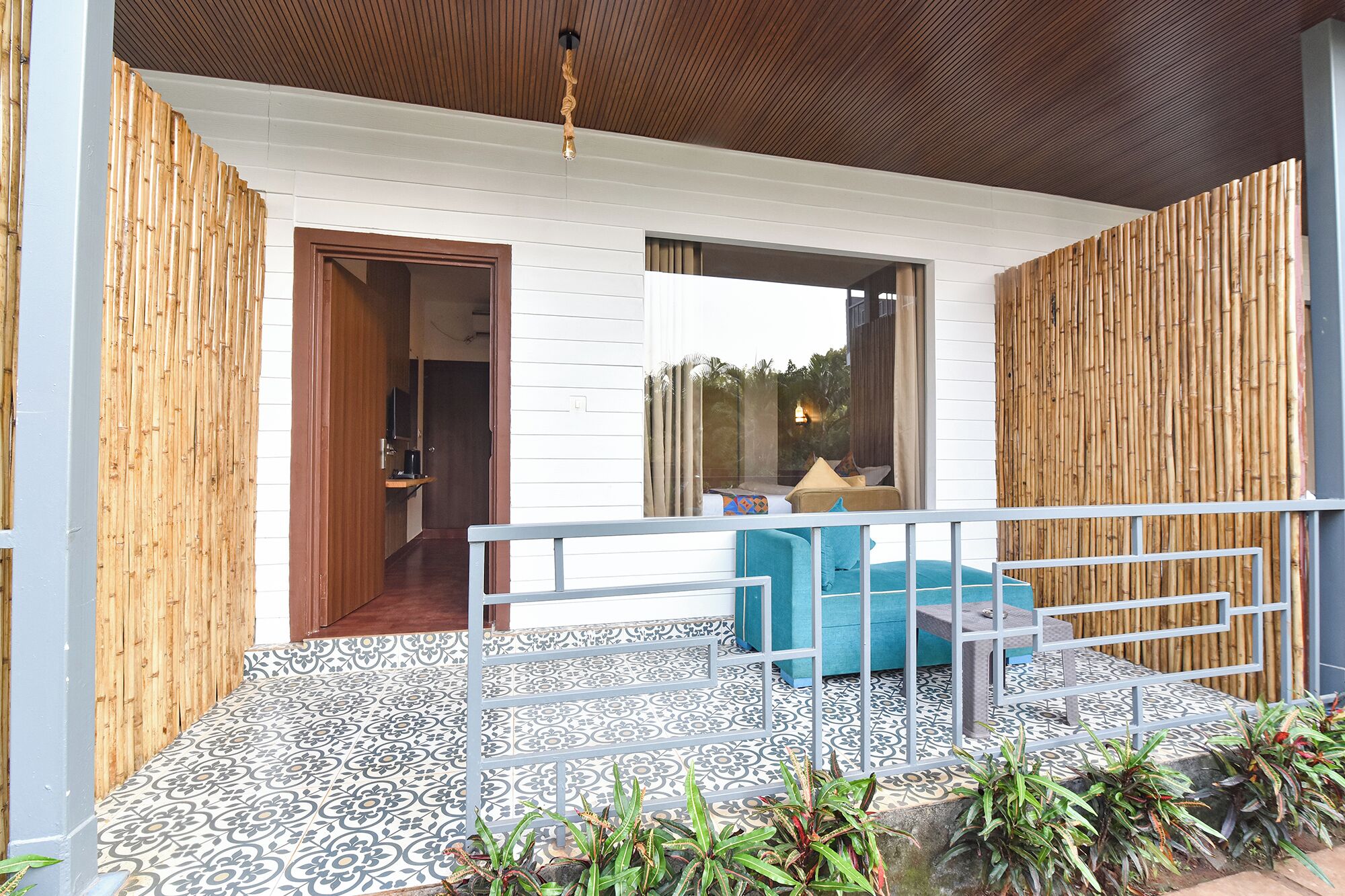 A covered outdoor seating area with a patterned floor, a wooden fence, and a view of the interior.