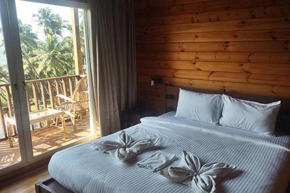 A wooden cabin room with a bed, a balcony with chairs, and a view of palm trees.