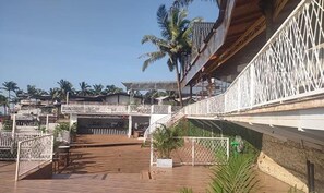 A wooden deck with a white railing, palm trees, and a view of a residential area with houses and a bridge. - Fabhotel Levels By The Sea (Vagator)