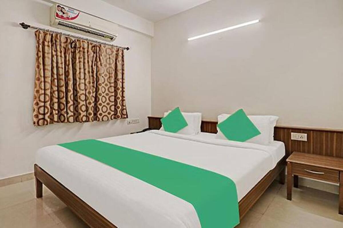 Quarto de hotel com cama, mesa de cabeceira, colcha verde, cortinas estampadas e unidade de ar condicionado.